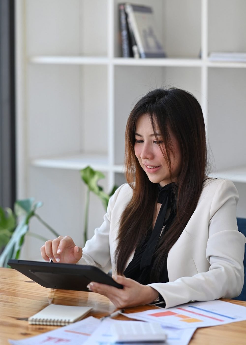 Businesswoman analyzing financial data on a tablet with financial documents spread on her desk.